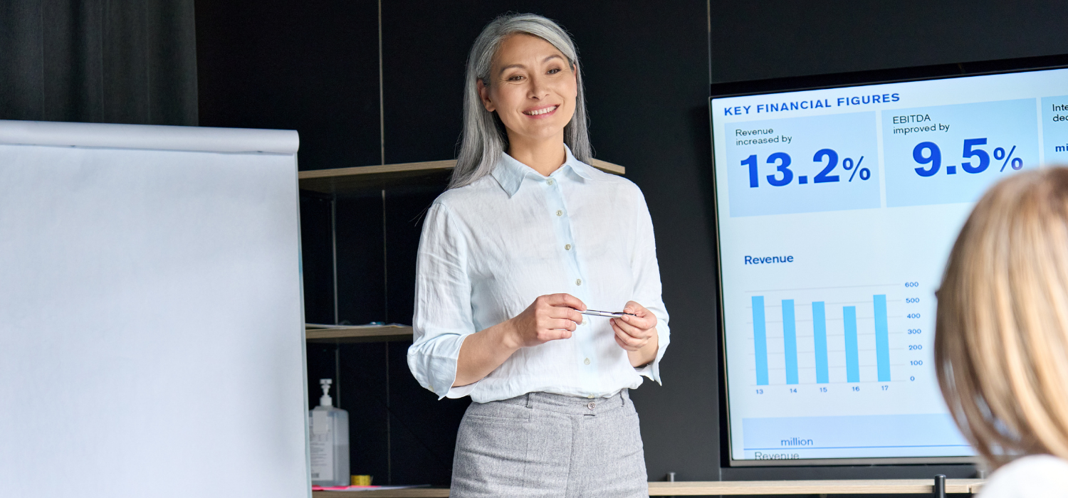 A female advertising manager with long ash silver-gray hair is explaining how hotel booking websites work to clients in a meeting room