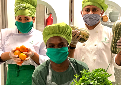 Three chefs in masks and white robes are holding up fresh produce for the camera.