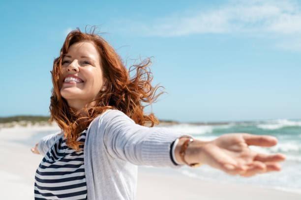 Mature woman enjoy breeze at beach Happy mature woman with arms outstretched feeling the breeze at beach. Beautiful middle aged woman with red hair and arms up dancing on beach in summer during holiday. Mid lady in casual feeling good and enjoying freedom with open hands at sea, copy space. good moods and energy stock pictures, royalty-free photos & images