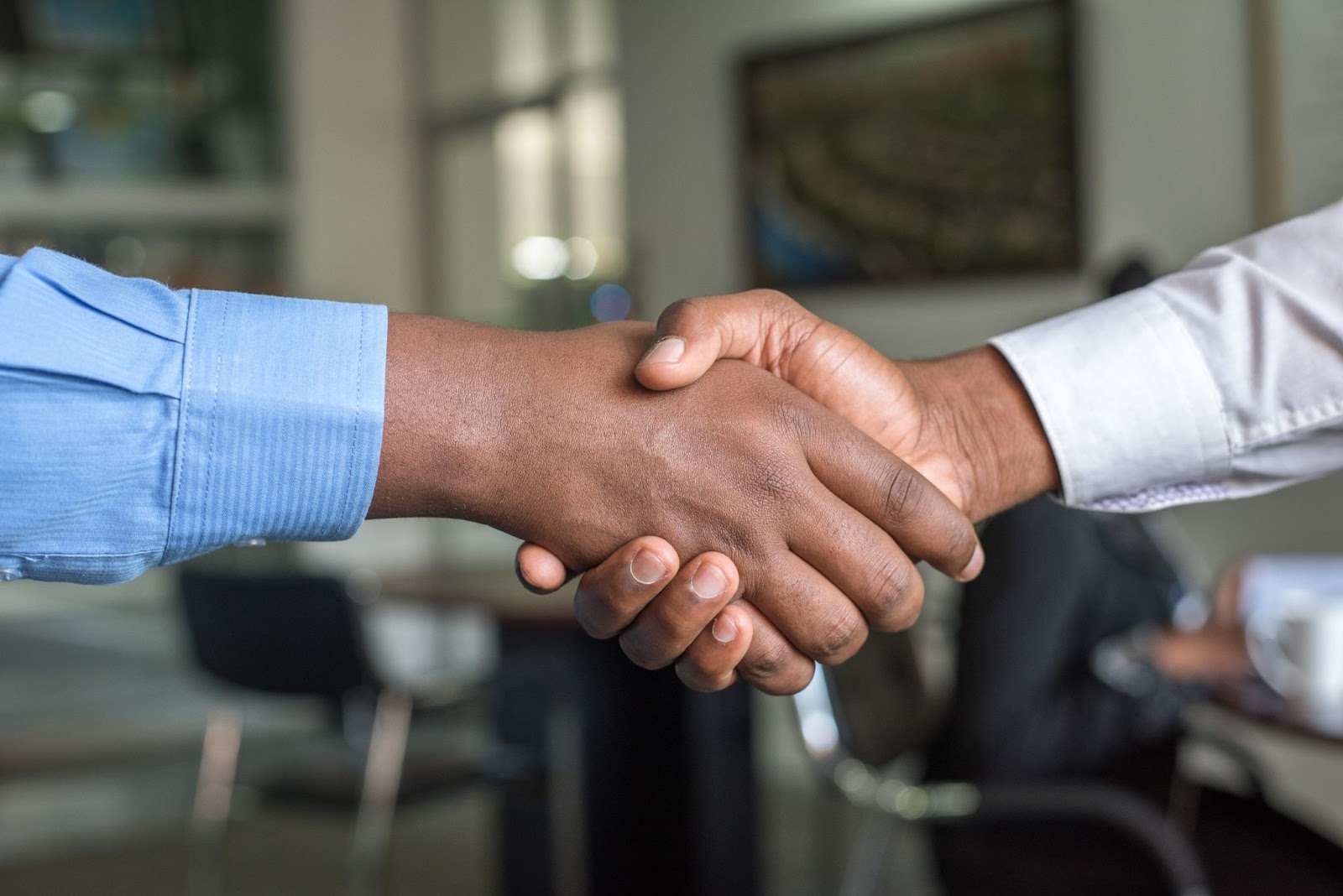 A imagem mostra em foco um aperto de mão entre dois homens negros. O que está localizado à esquerda utiliza camisa social azul, enquanto o da direita usa a mesma peça, mas branca. Ao fundo, desfocado, é possível reconhecer um ambiente que remete à um escritório de trabalho.