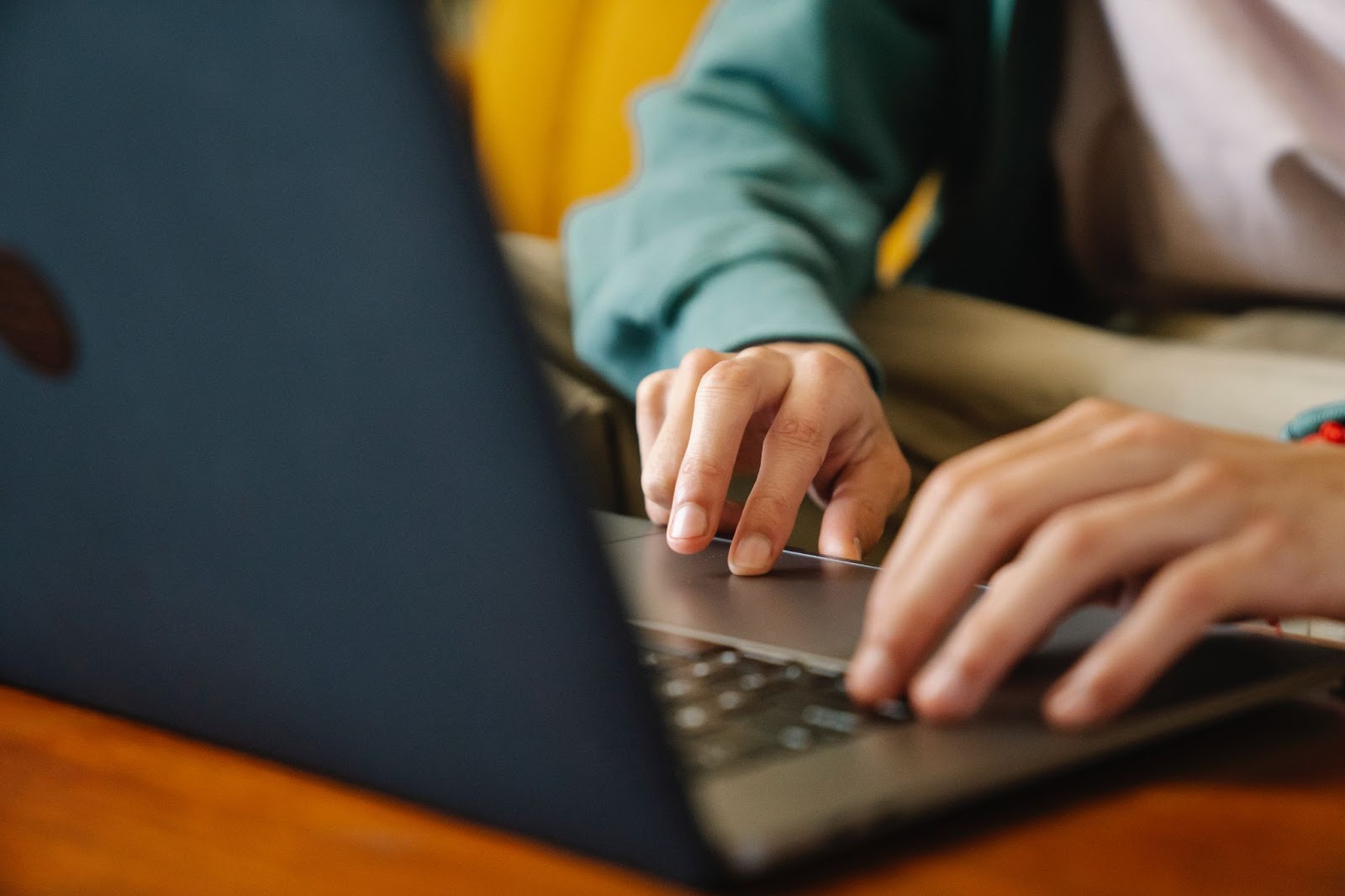 Imagem de um notebook está aberto sobre uma mesa de madeira. Em foco, estão duas mãos humanas que digitam em seu teclado.