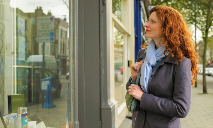 A woman looking through a storefront window and deciding if a new purchase is necessary