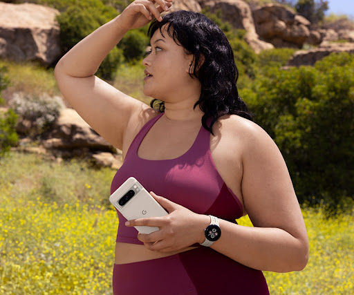 A person gazes around on a hiking trail. In their left hand is a Pixel 8 Pro in a Pixel 8 Pro Case. They also wear a Pixel smartwatch.