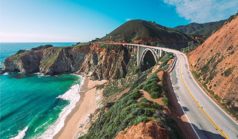 Aerial view of the Pacific Coast Highway 1 in California. There are mountains to the right, the pacific coast to the left, and a bridge up ahead.