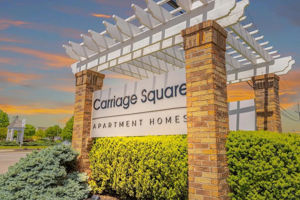 Entrance sign reading "Carriage Square Apartment Homes" flanked by brick columns and green shrubbery under a decorative pergola.