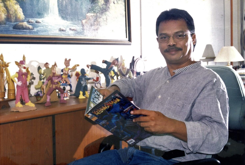 Photograph of Kamn Ismail sitting in a chair holding a magazine. He has brown skin, dark hair and mustache, and is smiling. Many action figures are on table behind him.