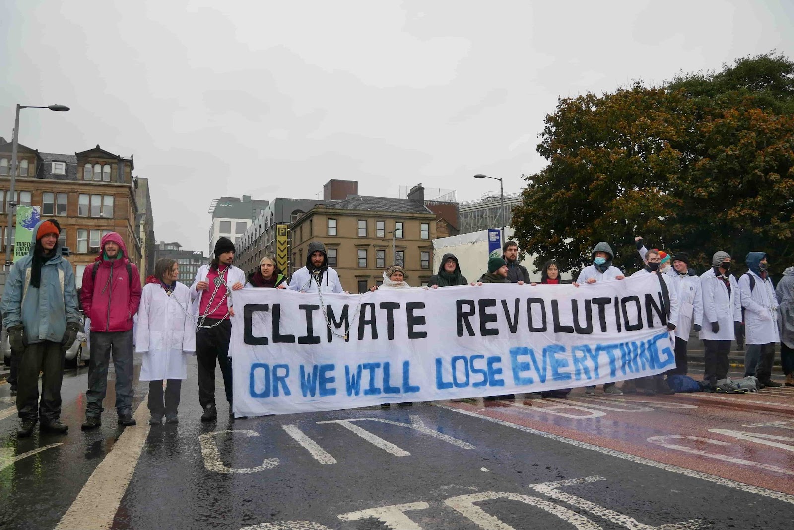 Scientists dressed in white coats stand in a long line across a road holding a sign which says 'climate Revolution or we will lose EVERYTHING'