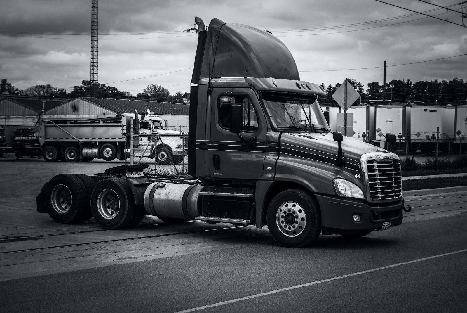 A black-and-white picture of a semi-trailer driving out of a lot filled with fleet semi-trucks in the background