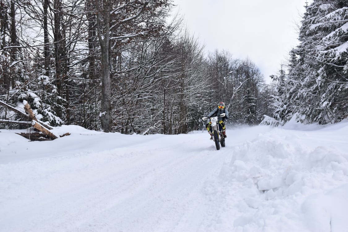 A motorcycle rider riding on a snow-covered road between trees on either side. Riding in the snow is unsafe and risky, more son in a remote forest area.