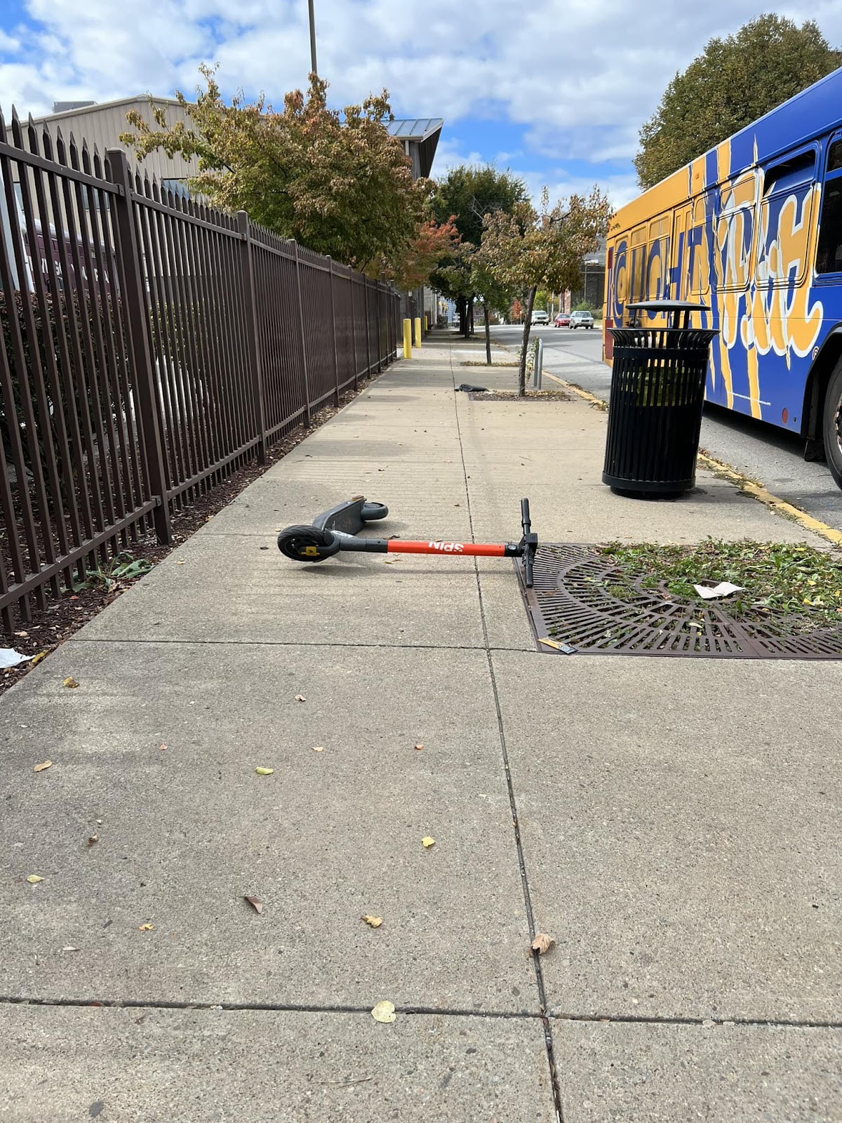 This is an image of a Spin scooter laying lengthwise across a sidewalk, sandwiched between a gate and a sewer grate, with a PRT bus pulling up to the curb.