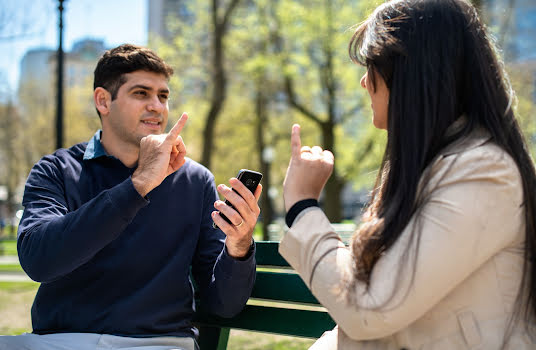 Um homem e uma mulher sentados num banco de jardim a falarem em linguagem gestual com a ajuda de um telemóvel.