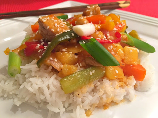 A mixture of pork, green onions, pineapple chunks and bell peppers on a bed of rice on a white plate with chop sticks.