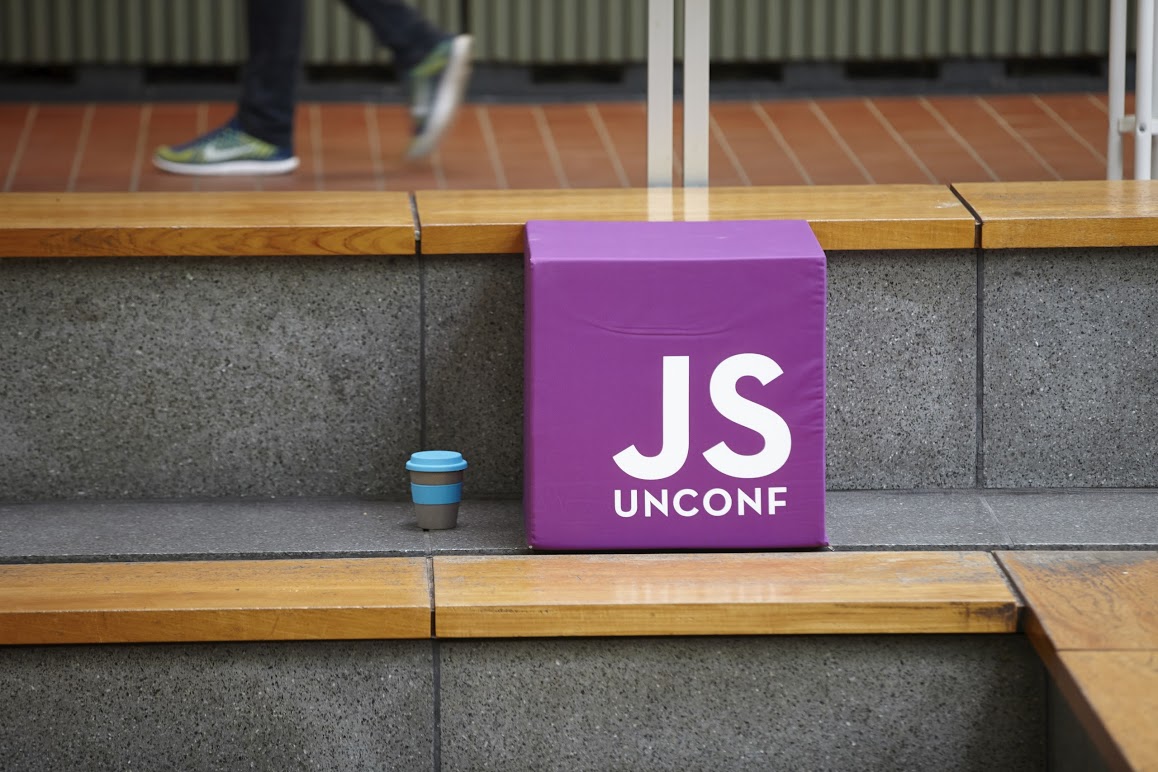 A photo of a large purple footstool with the JSUnconf logo next to a reusable JSUnconf coffee cup, with the walking feet of a conference attendee visible in the background