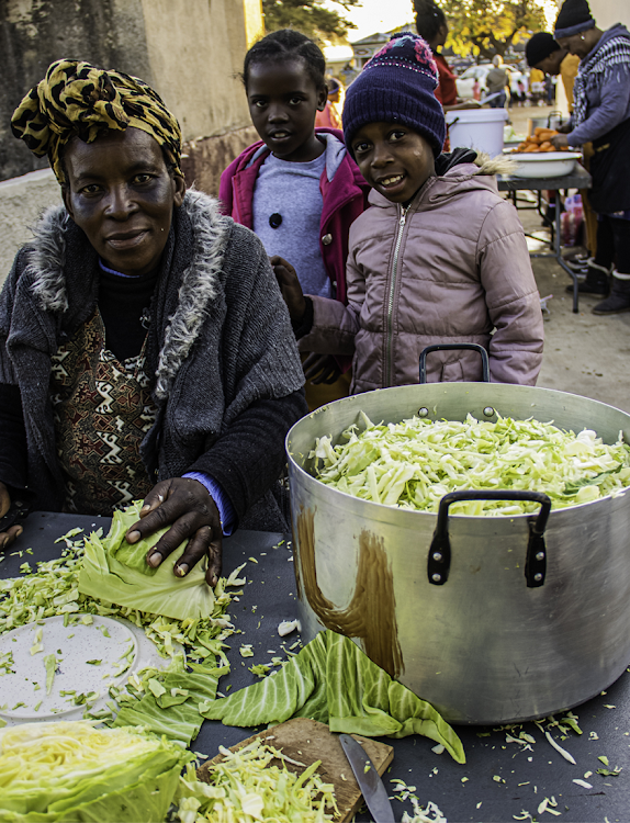 A relative chops cabbage (left), watched by youngsters Nokuthula Oratile (centre) and Ipileng Letado (right). In the background, sweet potatoes, carrots and other veggies are being prepared in large numbers.