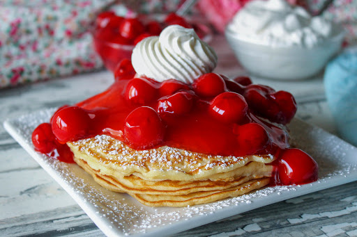 A stack of cherry vanilla pancakes with whipped cream on top.