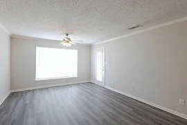 Living room with neutral flooring and walls, a ceiling fan, and a large window. There is also the door to access the patio 