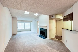 Empty apartment interior with carpeted living area, white walls, fireplace, and kitchen with brown cabinets.