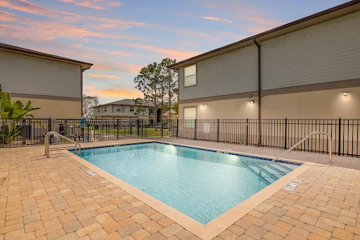 Pool area with clear blue water surrounded by a metal fence and flanked by apartment buildings at dusk.