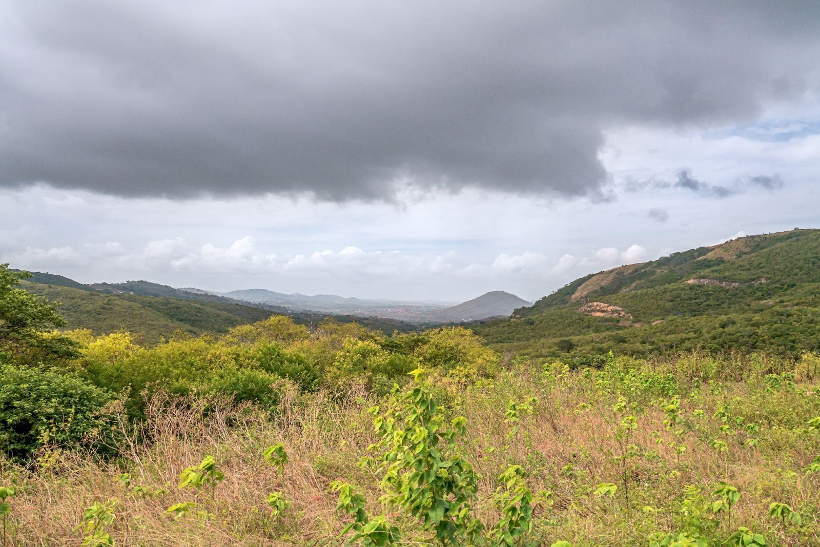 Paisagem das montanhas em Gravatá em tons de verde, com céu nublado e grande nuvem cinza.