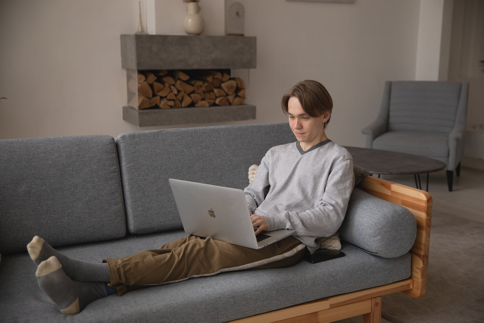 Boy in a grey sweater and brown pants lounging on a couch using his laptop