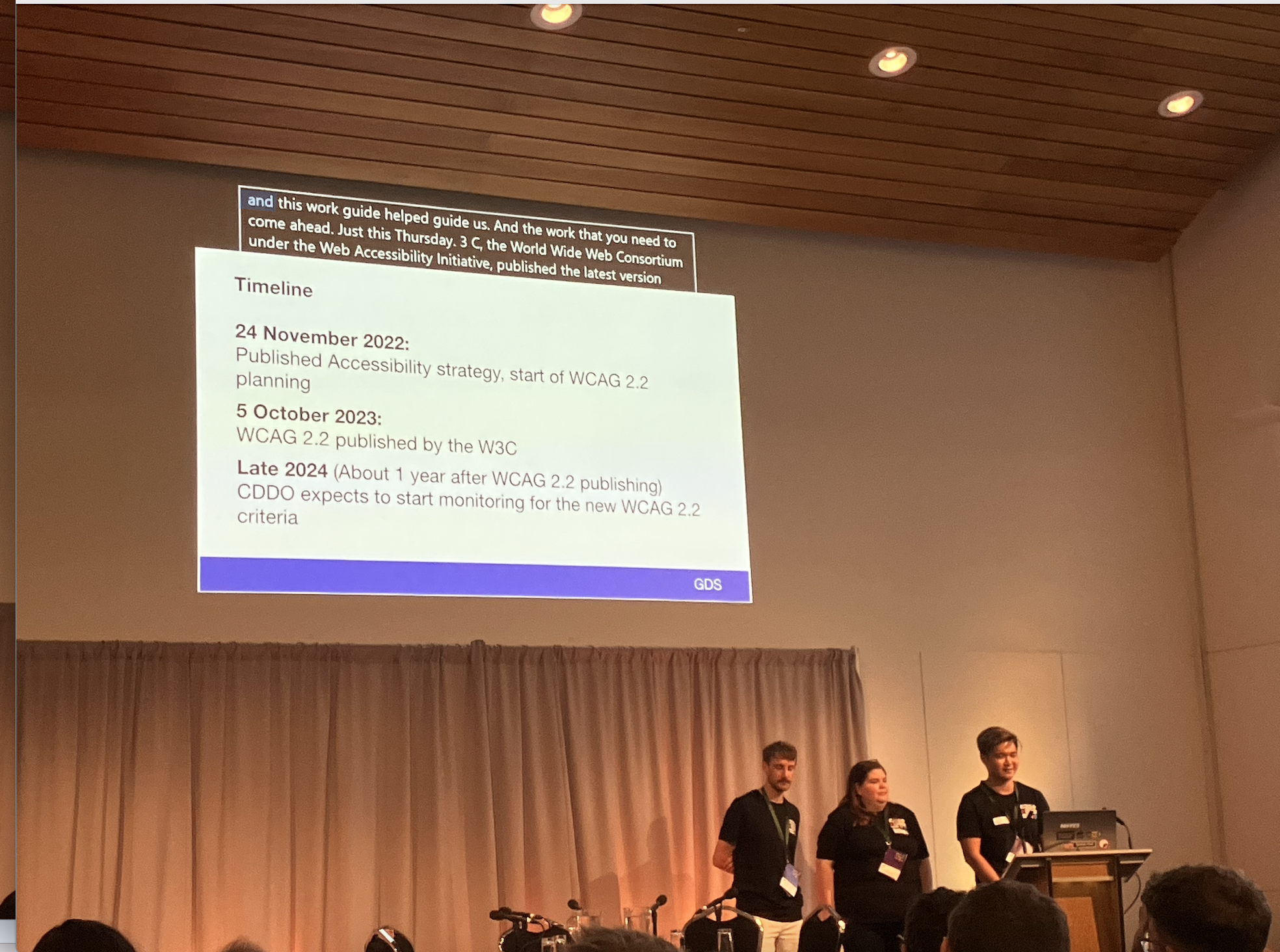 3 people on stage with screen saying "timeline - 24 November 2022: published accessibility startegy, start of WCAG 2.2 planning; 5 October 2023: WCAG 2.2 published by the W3C; late 2024 (about 1 year after WCAG 2.2 publishing) CDDO expects to start monitoring for the new WCAG 2.2 criteria)