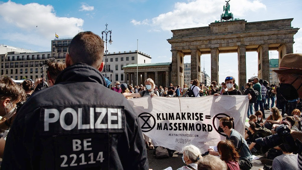 A policeman stands with his back to the camera. In front of him is a large group of rebels sitting down on some steps in a town square. Some people are holding a white banner. Most people have face masks on.