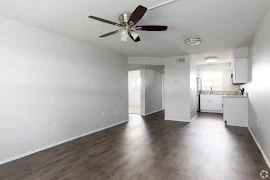 Livingroom with dark wood inspired flooring & neutral walls. In the background there is a kitchen, hallway, & bedroom. 