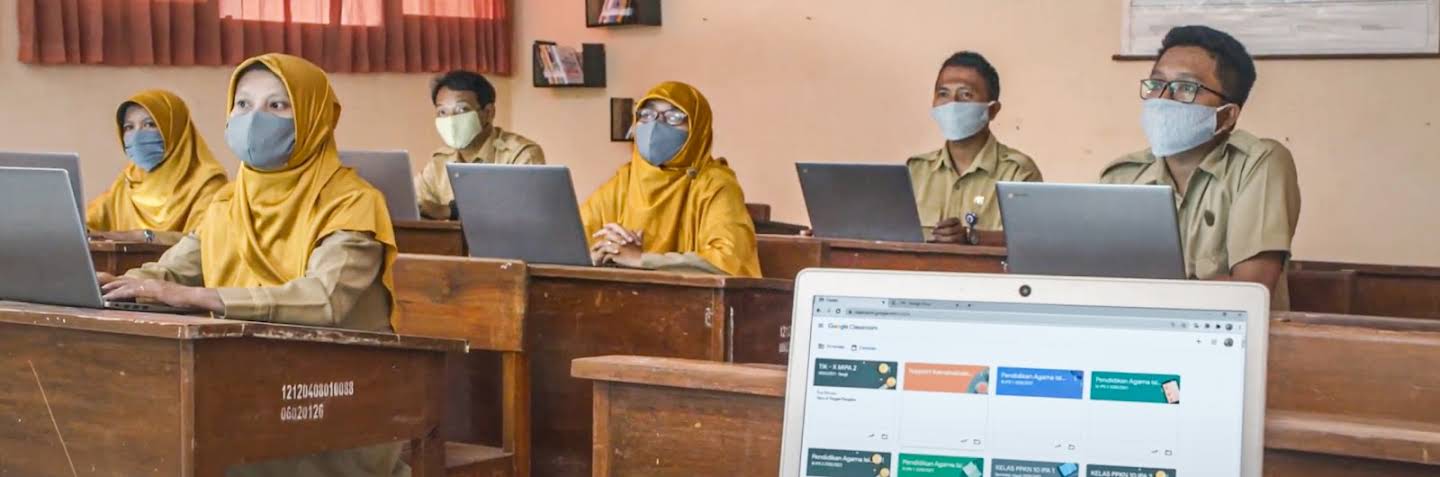 Tiga siswa yang mengenakan seragam sedang duduk di luar sebuah bangunan. Mereka semua mengenakan masker. Siswa di tengah menunjuk ke buku catatan yang diletakkan di atas keyboard laptop yang terbuka. Kedua siswa di sampingnya menonton dengan penuh perhatian.