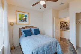 Model bedroom with blue comforter, plush carpet, light walls, wall decor, side lamp, ceiling fan, and view of entryway door