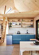 Tucked beneath the mezzanine, the kitchen — with its solid-larch above-counter shelving and drying rack, reclaimed travertine worktops and blue-green cabinets — forms part of the open-plan living area.