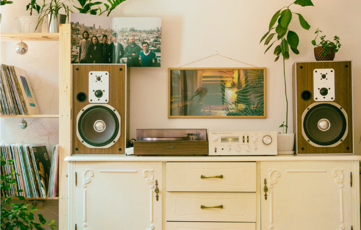 A white dresser with a record player, speakers, and mixer on top of it. There is a bookcase beside it, filled with vinyl records. A picture is hung on the wall and houseplants decorate the space.