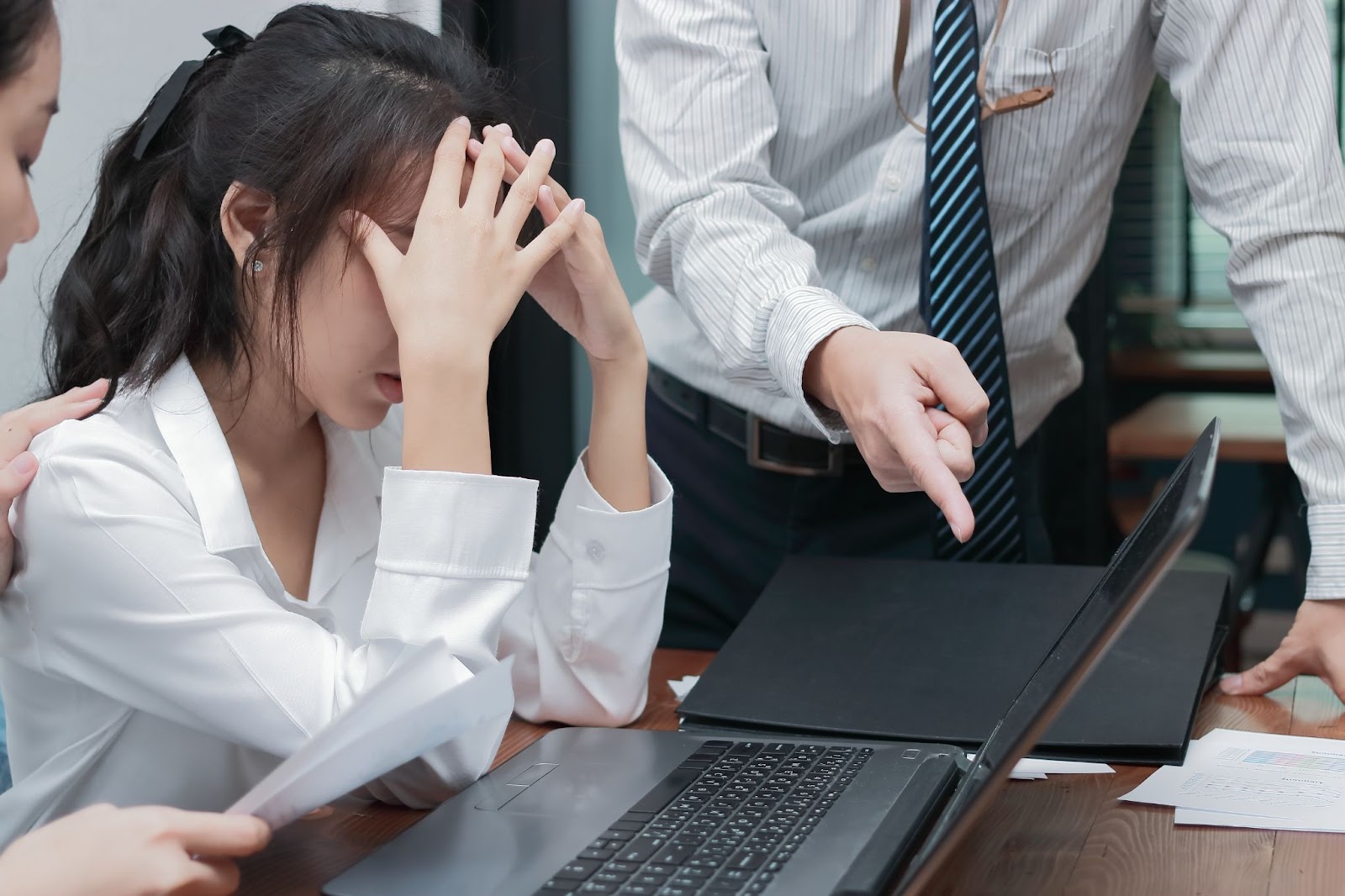 Asian businesswoman showing stressed face covering her face and man's hand pointing at laptop.