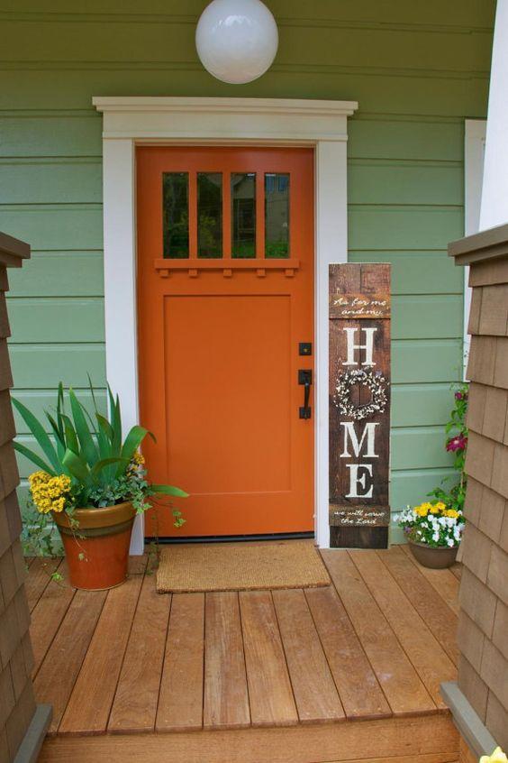 Fachada de casa com porta laranja, parede verde, deck de madeira e vasos de planta decorativos.