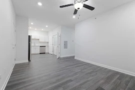 Modern apartment interior with white walls, gray flooring, ceiling fan, and kitchen appliances.