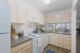 Model kitchen with light wooden cabinets, gray countertops, white appliances, stainless steel sink, and decorative accents