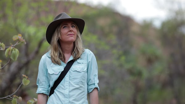 Dr. Emily Vink, who provides healthcare through Prescript, stands looking at the sky. She has trees around her and wears a wide brim hat.