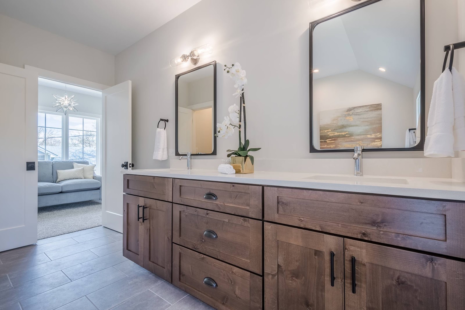 Natural Wood dual sink vanity with center bank of drawers and white quartz countertops. Two individual metal framed mirrors mounted above each sink with a modern vintage bulb fixture overhead.