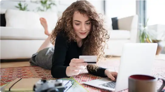 Uma mulher com cabelos longos e cacheados faz compras on-line. Ela digita um número de cartão de crédito enquanto está deitada de bruços no chão.