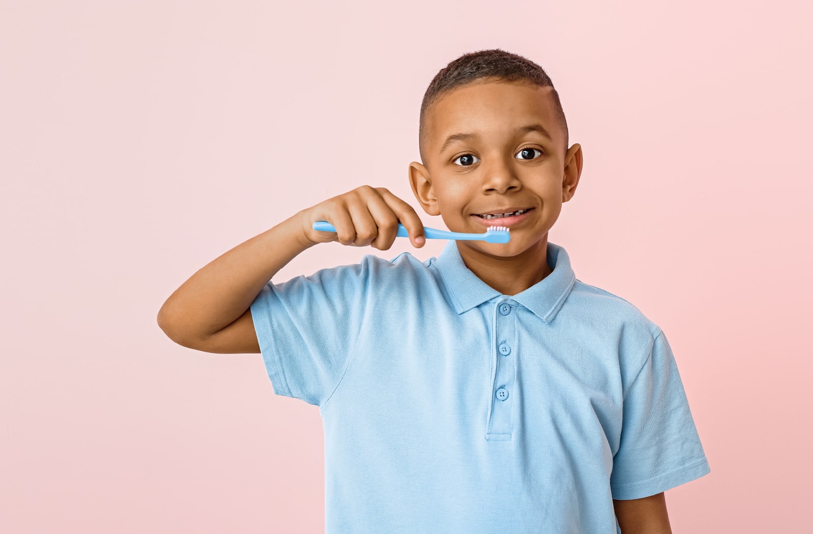 A young boy wearing a blue shirt, standing against a pink background, holds a toothbrush up to his mouth