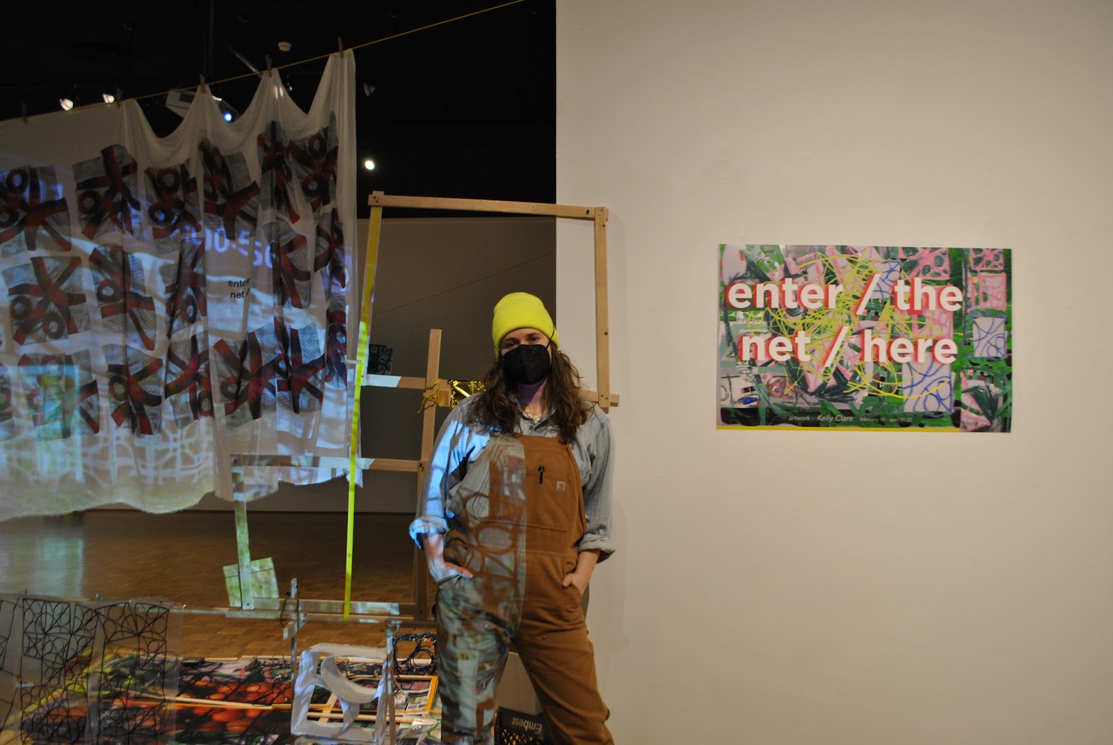 Image: Kelly Clare, enter / the net / here, 2022, Edna Carlsten Gallery. Clare stands center frame near an exhibition graphic. Behind the artist are hanging textiles, projected images, and sculptural elements in the gallery space. Image courtesy of the artist.