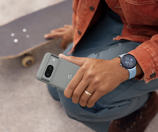 A person kneels beside their skateboard. In their left hand is a Pixel 8 in a Pixel 8 Case. They also wear a Pixel smartwatch.