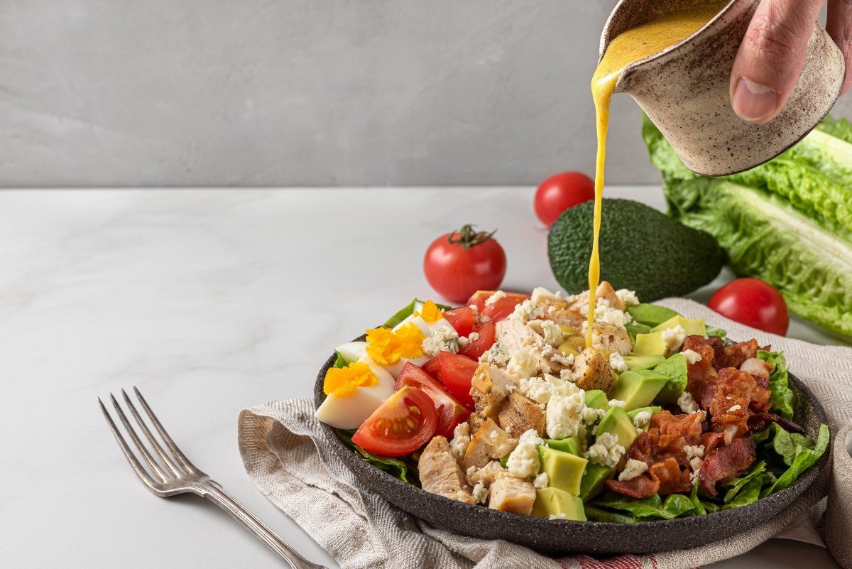 A hand pouring salad dressing into cobb salad with chicken