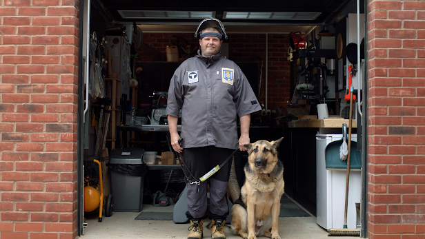A blind man stands with a guide dog in front of a garage