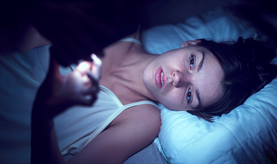 A young Caucasian woman laying in bed in the dark and using her phone instead of sleeping. This photo represents how we tend to use our phone late into the evening and have sleep problems as a result. 