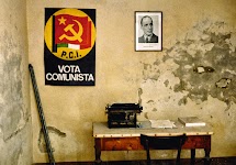 sober kamertje met vochtvlekken in de muur, waarop een foto van een man en een communistisch affiche, en een kleine tafel met typemachine en stoel