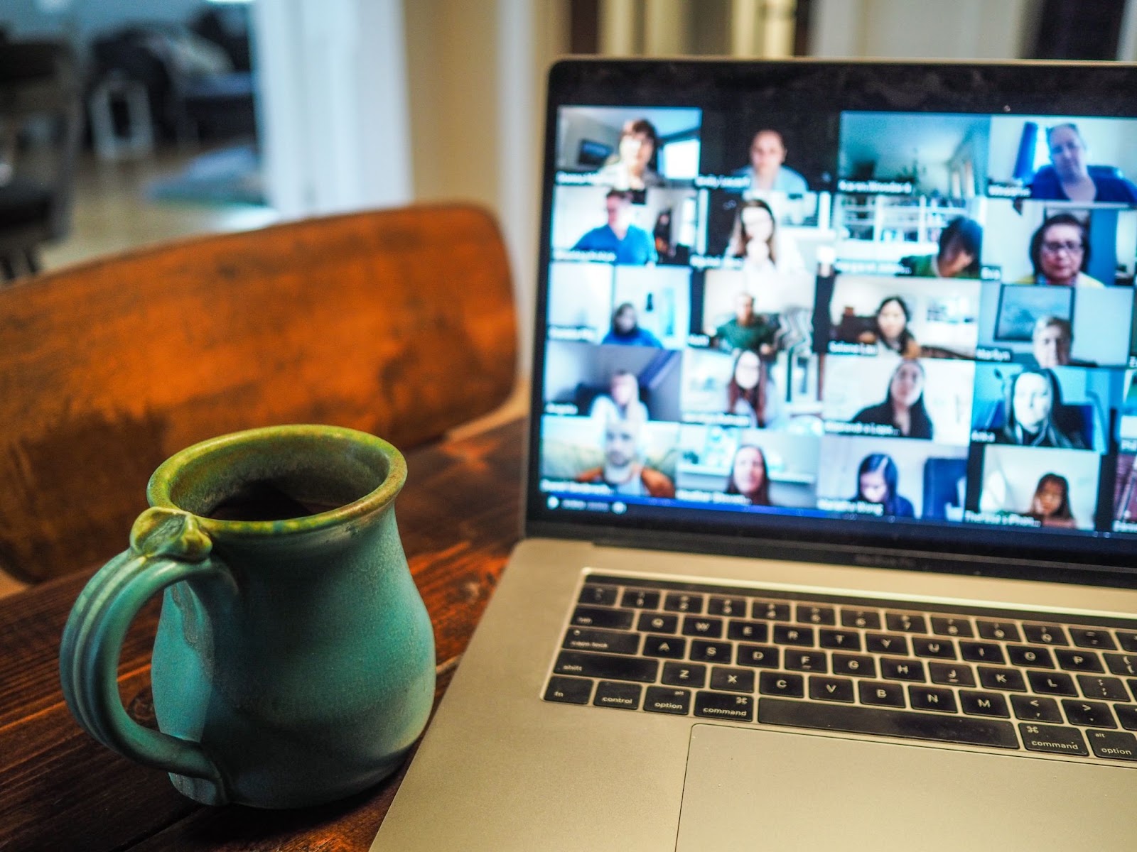 A imagem mostra um notebook prata aberto numa videoconferência,  como se fosse uma reunião em inglês, onde é possível ver ao menos vinte imagens de participantes. Ao lado esquerdo dele, há uma caneca azul esverdeada. O notebook e a caneca estão sobre uma mesa de madeira.