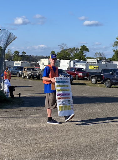 Event Worker wearing an orange safety vest holds informational sign while directing traffic on a sunny day