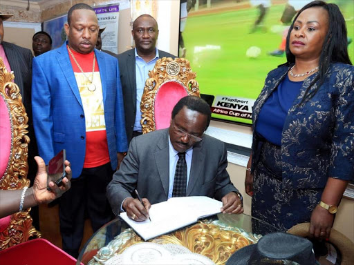 Wiper leader Kalonzo Musyoka signs a visitors' book during the commemoration of Nairobi governor Mike Sonko’s father's anniversary in Machakos on Saturday. Former MP Wavinya Ndeti is standing right. /GEORGE OWITI