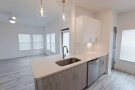 Kitchen island with stainless steel sink, pendant lighting, & white stone counters. In the back is a view of the living room