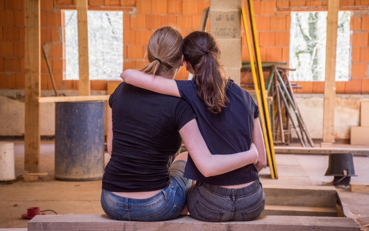 Two women wearing black tops and jeans embrace each other, looking at their home in the middle of a full remodel